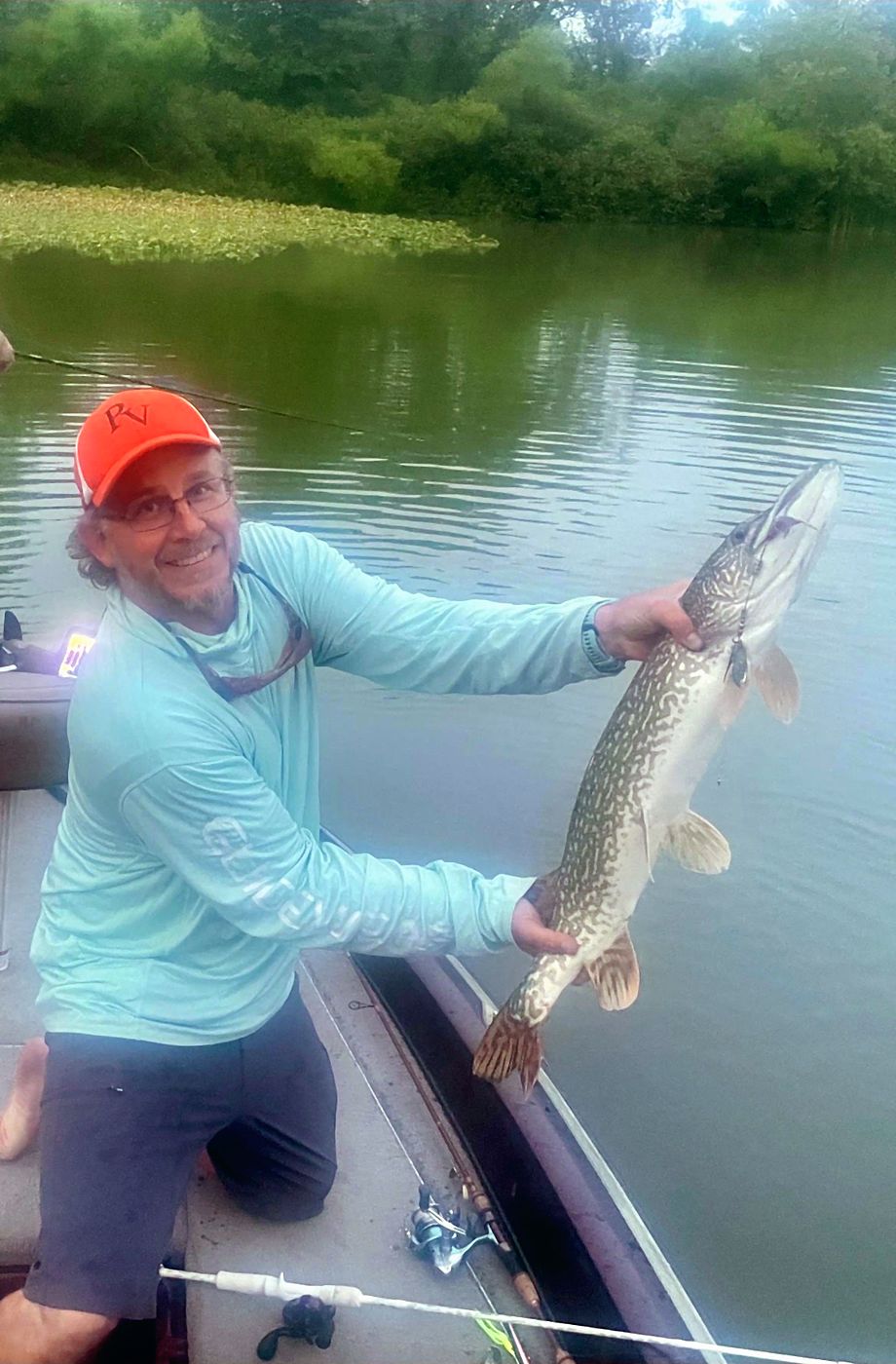 A northern pike held out over the water from a boat, lily pads and a wooded bank behind.