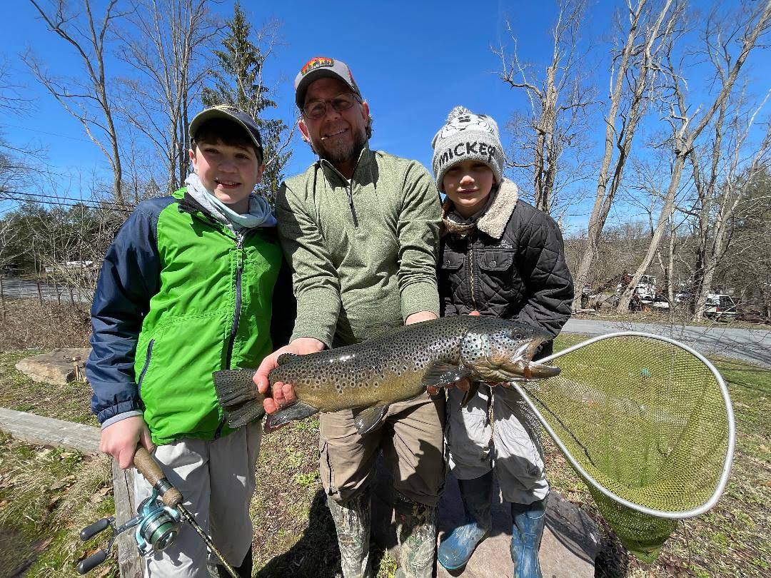 Three anglers on a pond bank in early spring holding a 25 inch brown trout, landing net and spinning rod beside them.