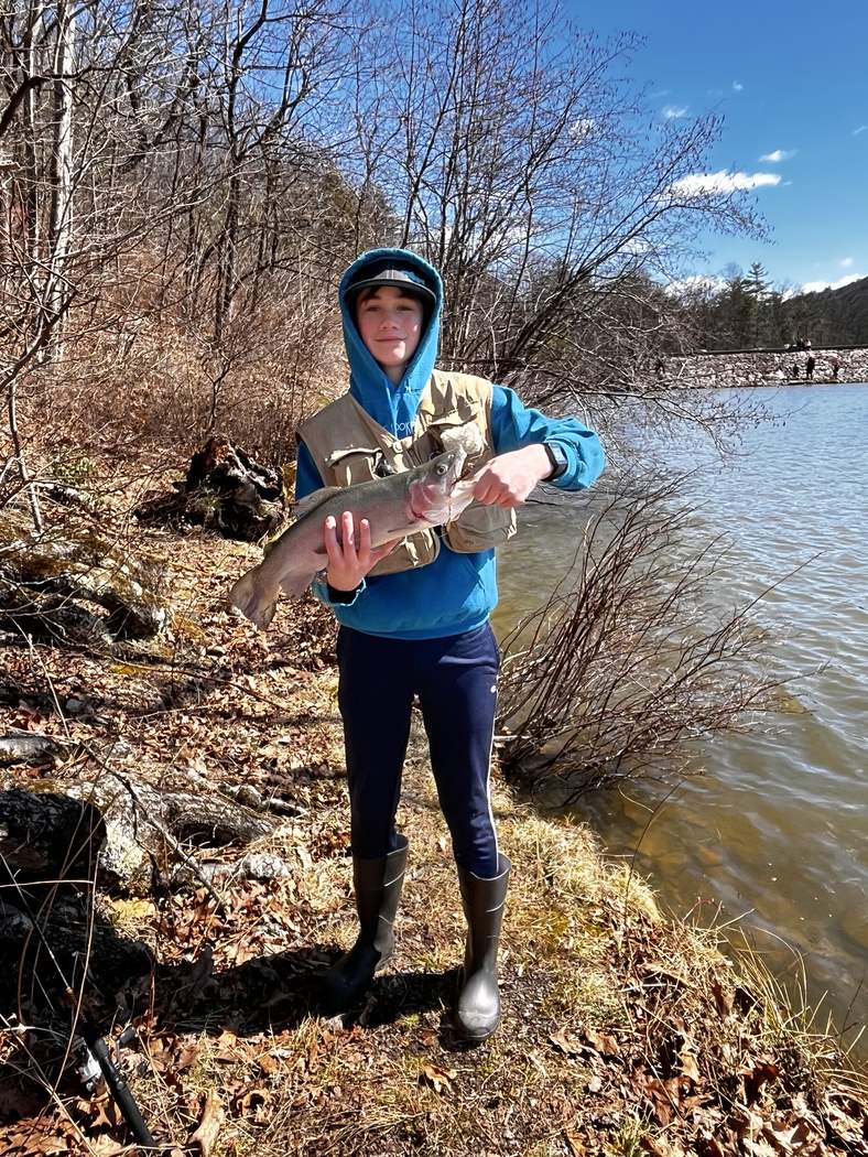 A rainbow trout held at the shoreline of a lake in early spring, bare timber on the far bank.