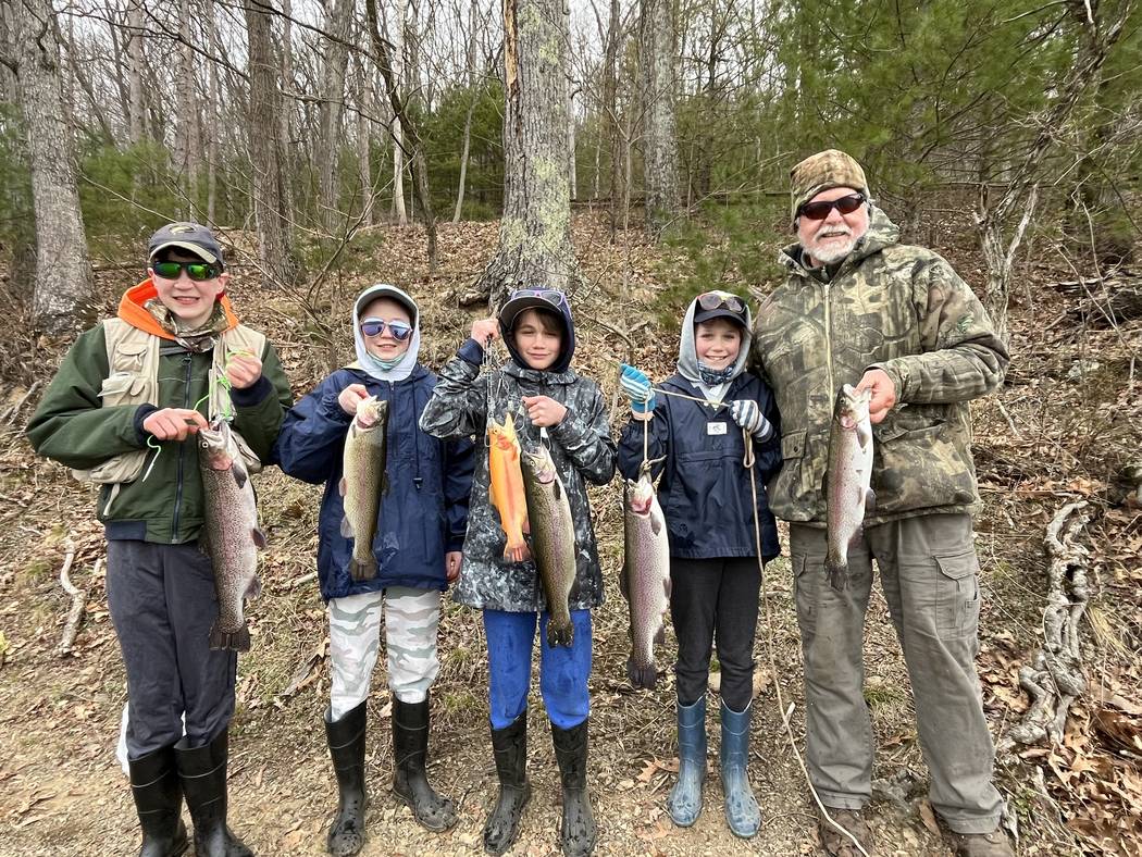Four young anglers and one adult lined up along a wooded bank, each holding a trout.
